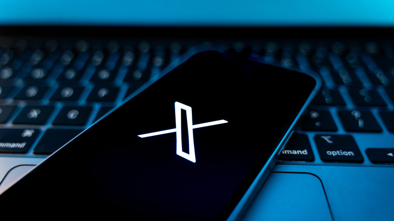 Photo of a smartphone screen showing the X social media logo. The phone has been placed on a laptop keyboard and is lit by blue screen glow.