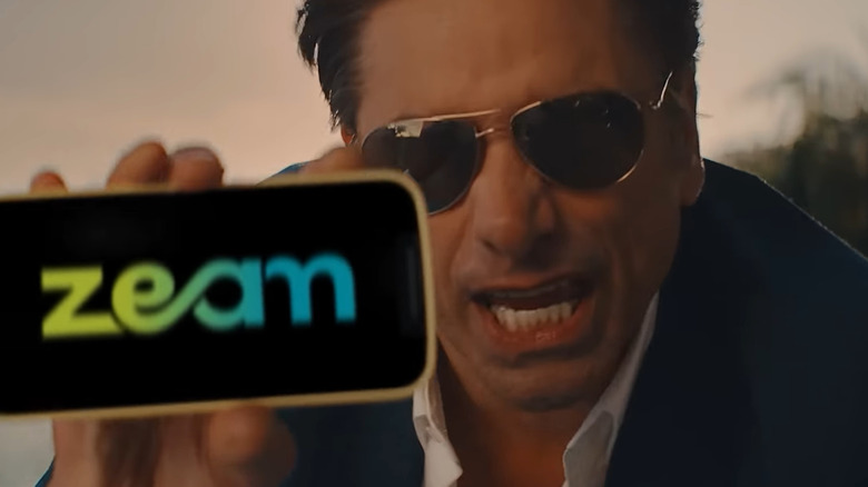 John Stamos in sunglasses shows off his phone with the Zeam logo in a commercial for Zeam