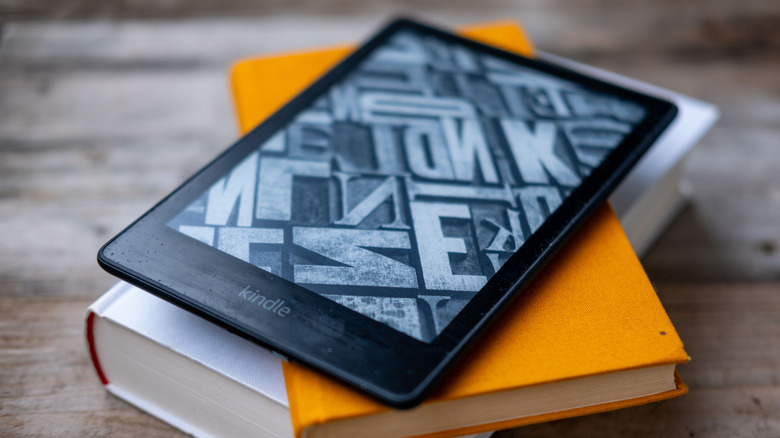 A kindle sits atop a stack of hardcover books atop of a wooden table.