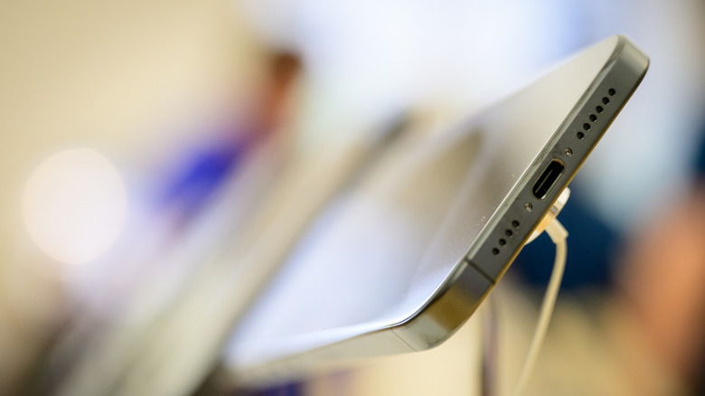 Close-up photo of the USB-C port on an in-store iPhone model. Only the bottom edge of the phone is in focus; the rest of the photo is blurred.