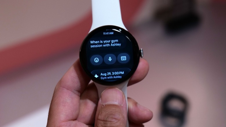 A Pixel Watch 5 held up with a message onscreen asking about the owner's upcoming gym session. A Gemini-suggested reply below offers a date and time.